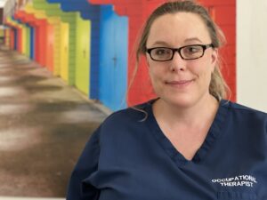 Paula Thompson wearing a blue tunic that says 'Occupational Therapist' on, standing in front of a row of coloured doors