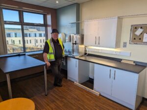 Construction worker standing n kitchen with a window behind him. Kitchen units look dated.