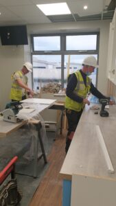 Two men fitting a kitchen, one is moving a box and the other is using a cordless drill