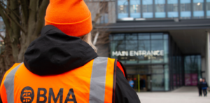 Back of junior doctor wearing hi viz vest and orange hat looking at Hull Royal Infirmary main entrance