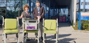 Woman and man standing outside hospital main entrance behind three new green wheelchairs