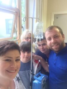 Zowie in hospital with a drip stand and members of her family around including her mum, two sons and partner. Zowie's hair has begun to grow back.