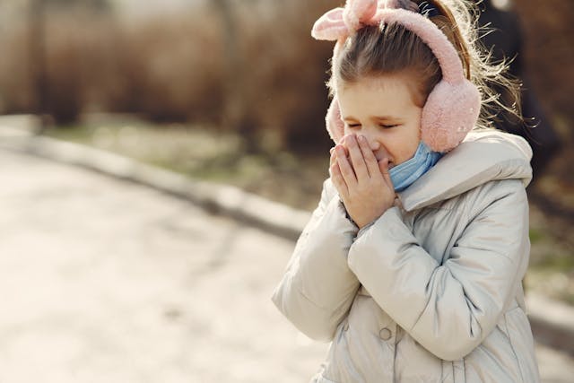 A child wearing pink ear muffs and a winter coat sneezes, holding her hands over her nose